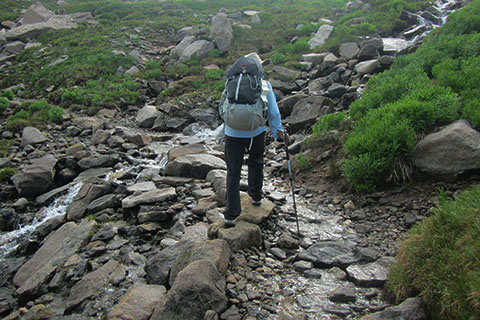 Hiker crossing another creek on rocks as the creek spills onto the trail